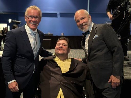 Un homme entre deux hommes en veston, devant un décor de conférence.
