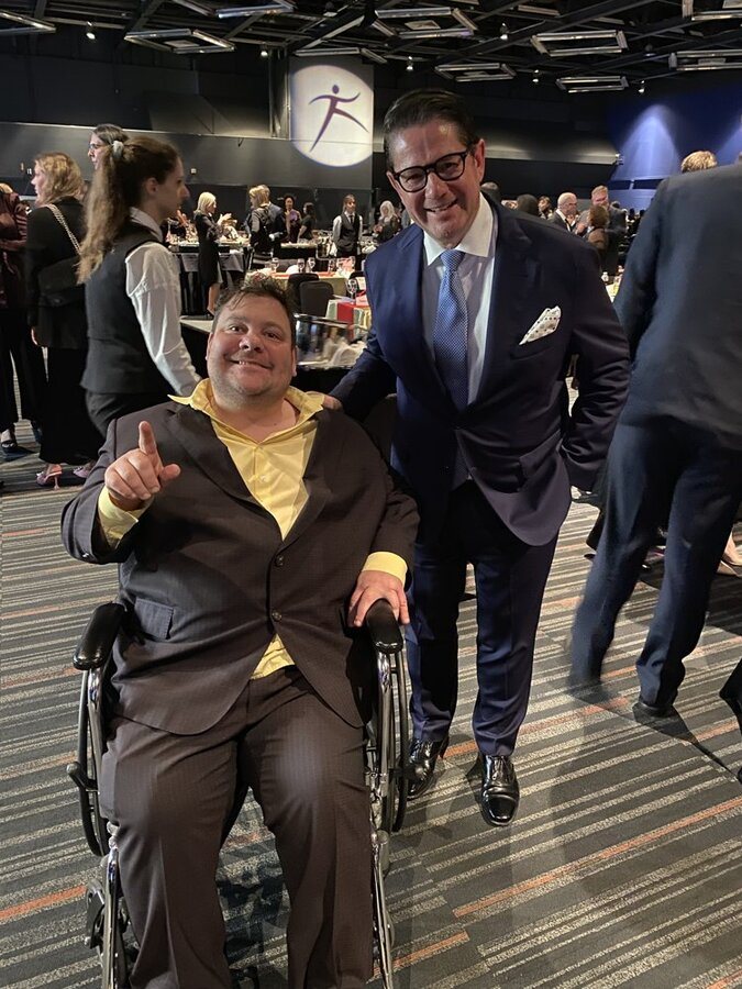 Dans un grand événement, un homme en veston se tient à côté d’une personne en fauteuil roulant.