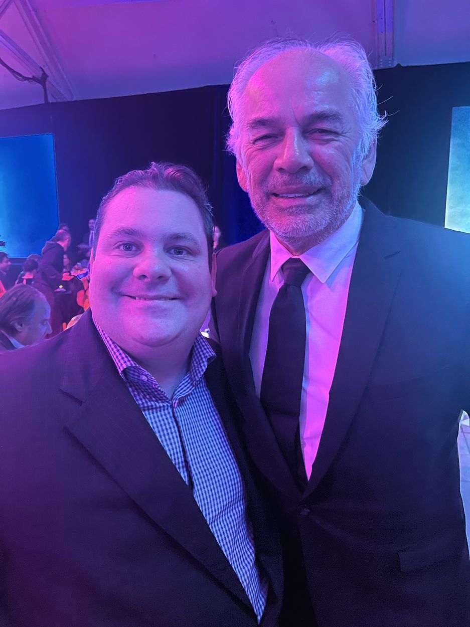 Deux hommes souriants posent lors d’une soirée, sous un éclairage violet.